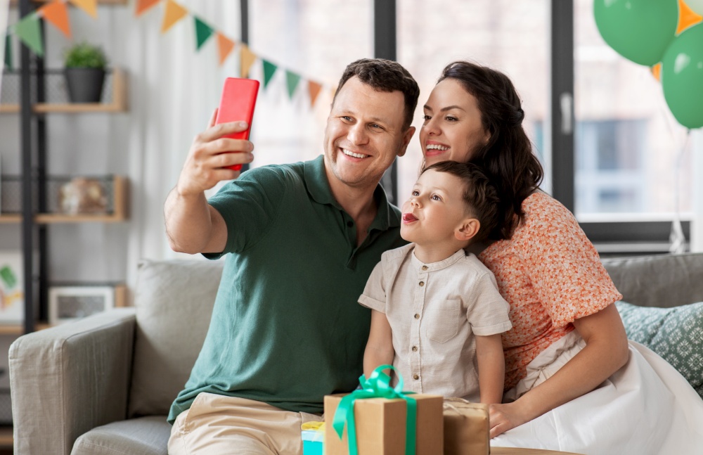 family, holidays and people concept - portrait of happy mother, father and little son with gifts and smartphone taking selfie on birthday at home party. happy family taking selfie on birthday at home