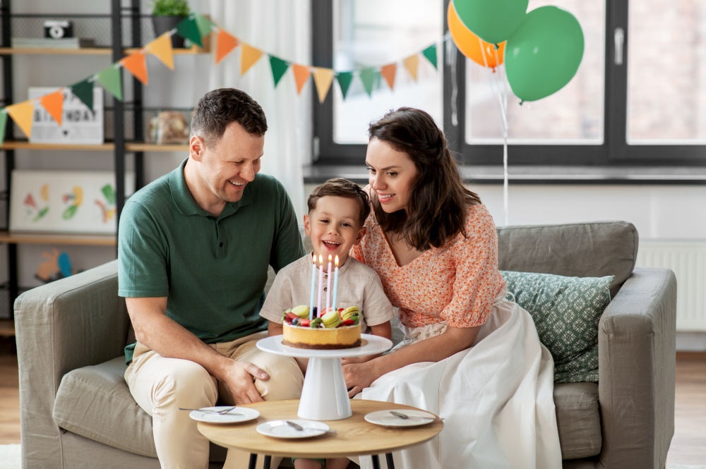 family, holidays and people concept - portrait of happy mother, father and little son with four candles burning on birthday cake sitting on sofa at home party. happy family with birthday cake at home
