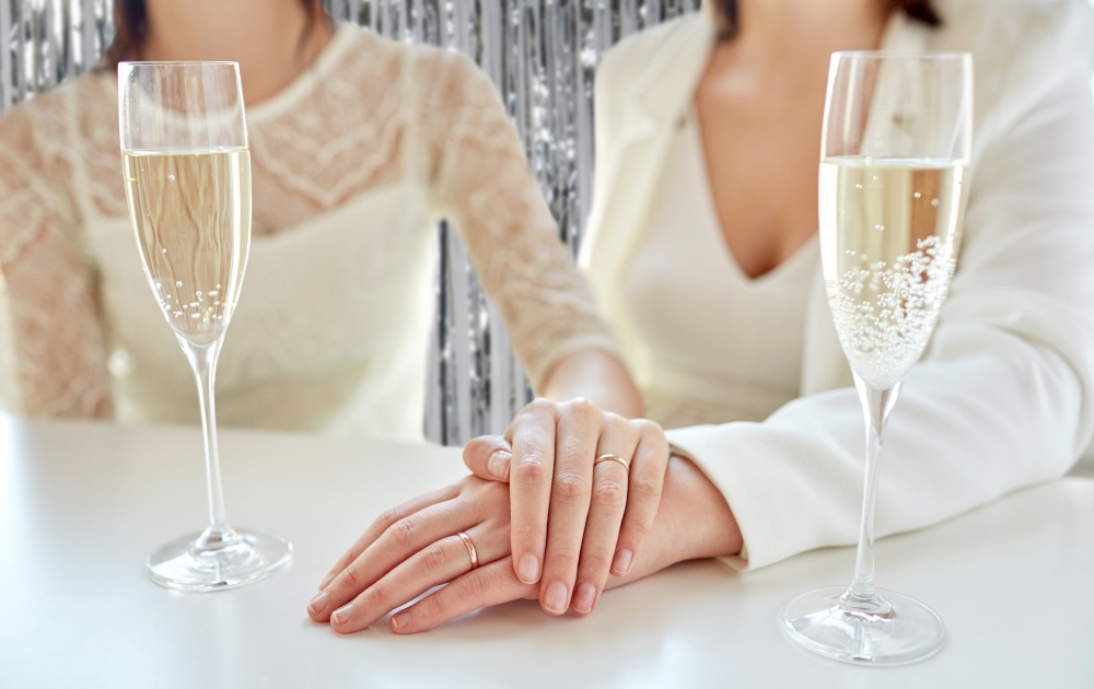 homosexuality, same-sex marriage and lgbt concept - close up of happy married lesbian couple hands with champagne glasses and wedding rings over foil party curtain on background. close up of lesbian couple with champagne glasses