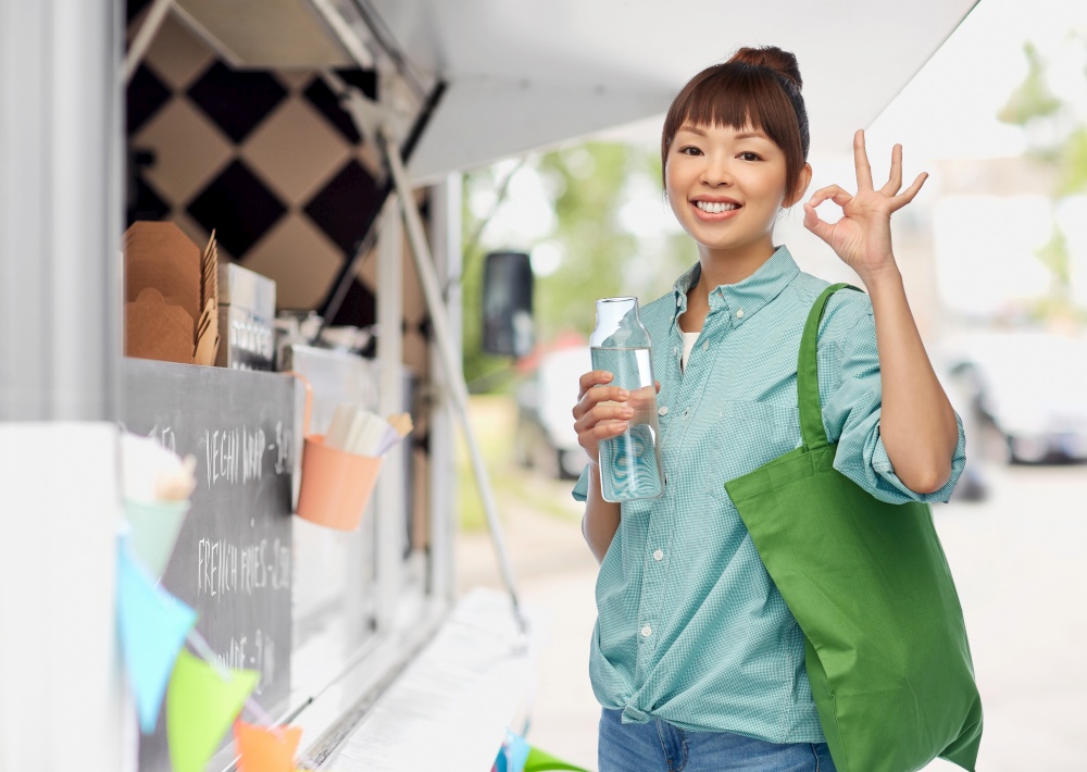 sustainability, eco living and people concept - portrait of happy smiling young asian woman with green reusable shopping bag and glass bottle of water showing ok gesture over food truck background. woman with shopping bag and bottle over food truck