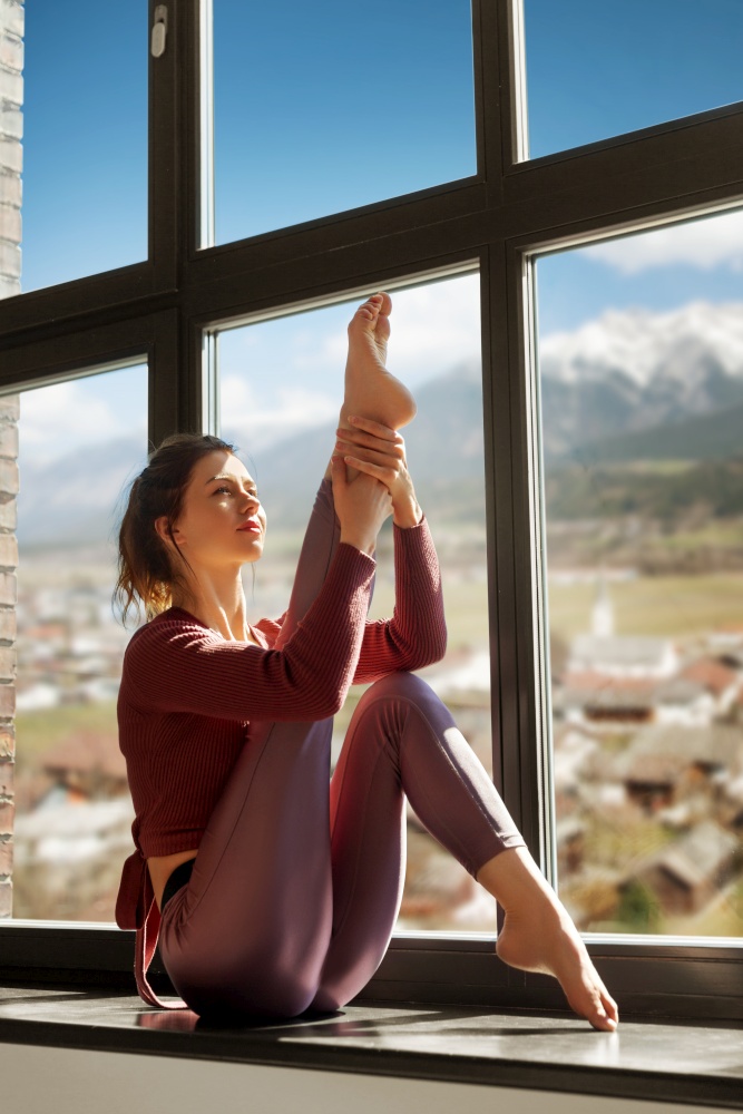 fitness, sport and healthy lifestyle concept - woman doing yoga exercise on window sill at studio over suburban view and alps mountains on background. woman doing yoga exercise on window sill at studio