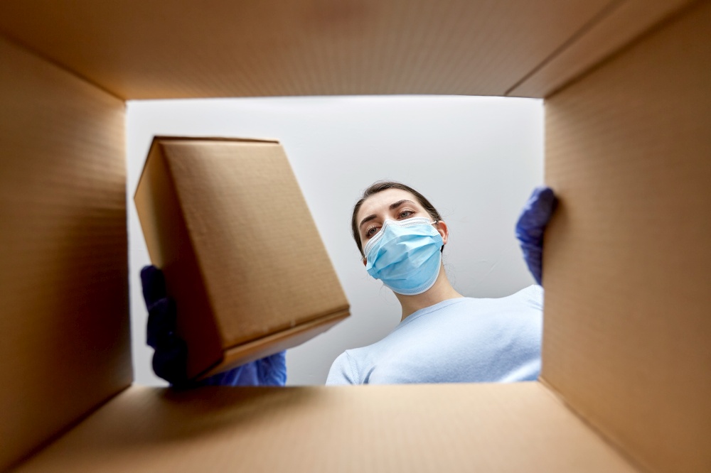 home delivery, shipping and pandemic concept - woman in protective medical mask and gloves opening parcel, looking inside and taking box out. woman in mask taking box from parcel