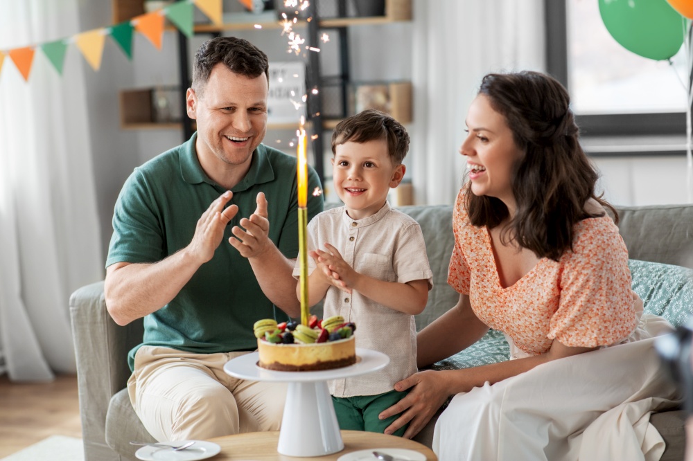 family, holidays and people concept - portrait of happy mother, father and little son with firework candle burning on birthday cake sitting on sofa at home party. happy family with birthday cake at home