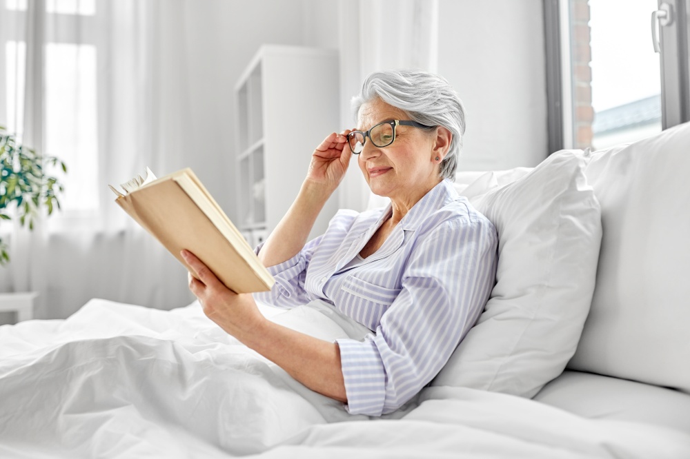old age, leisure and people concept - happy smiling senior woman in glasses reading book in bed at home bedroom. old woman in glasses reading book in bed at home