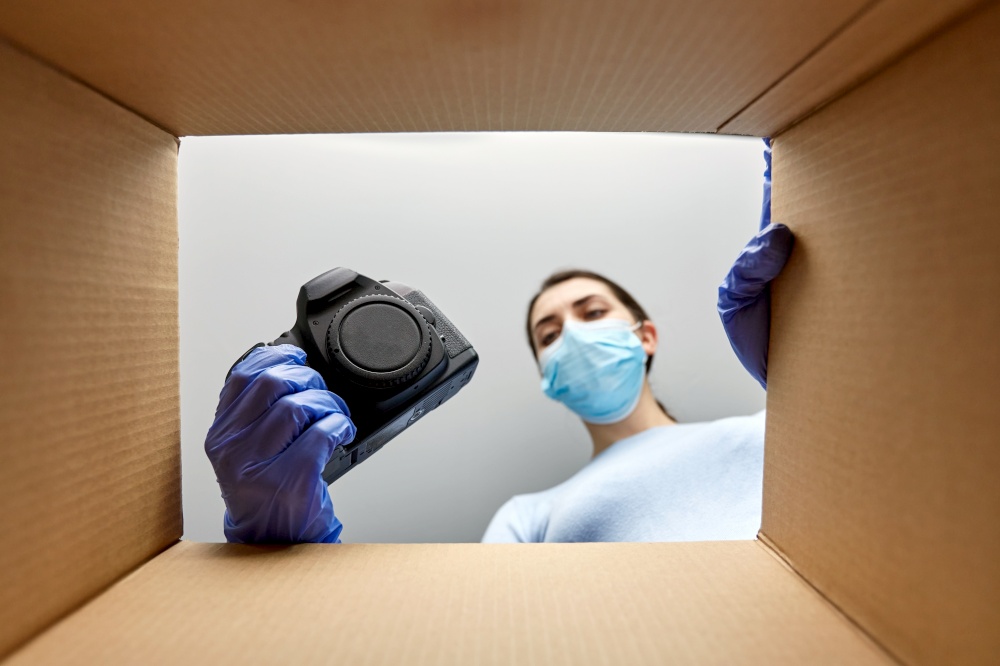 delivery, shipping and pandemic concept - woman in protective medical mask and gloves packing digital camera and lens into parcel box with foam peanuts. woman in mask packing camera into parcel box