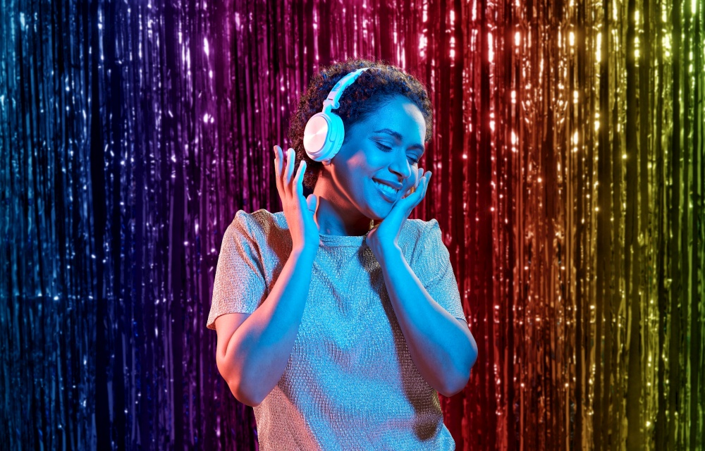 nightlife, technology and people concept - happy young african american woman in headphones listening to music and dancing at party in neon lights over rainbow foil curtain background. woman in headphones listening to music at party