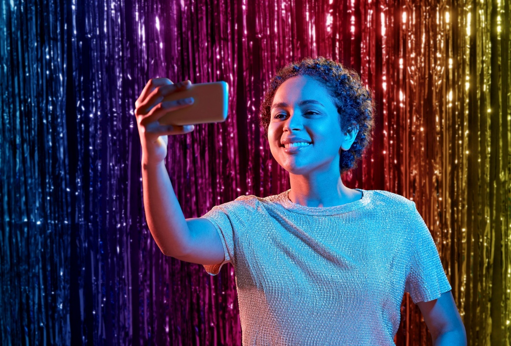 nightlife, technology and people concept - happy young african american woman taking selfie with smartphone at party in neon lights over rainbow foil curtain background. woman taking selfie with smartphone at party