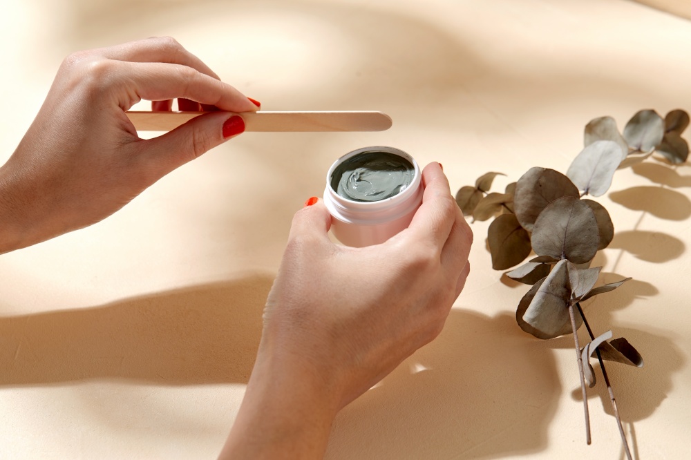 beauty, cosmetics and bodycare concept - hands holding jar of blue cosmetic clay mask and wooden spatula over branch of eucalyptus populus on beige background. hands holding jar of blue cosmetic clay mask