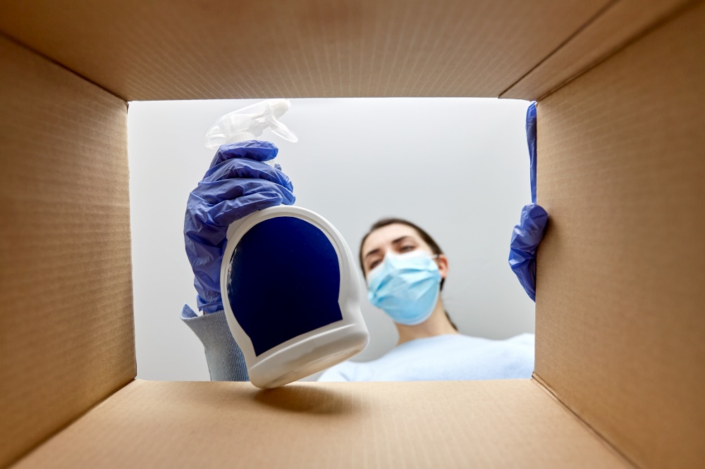 home delivery, shipping and pandemic concept - woman in protective medical mask and gloves opening parcel box, looking inside and taking cleaning supplies out. woman in mask taking cleaning supplies from box