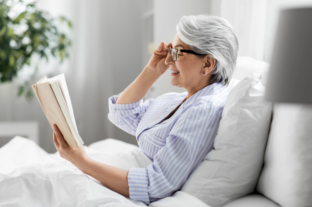 technology, old age and people concept - happy smiling senior woman in glasses reading book in bed at home bedroom. old woman in glasses reading book in bed at home