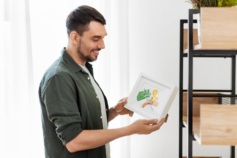 home improvement, decoration and people concept - happy smiling man placing watercolor picture in frame to shelf. man decorating home with picture in frame