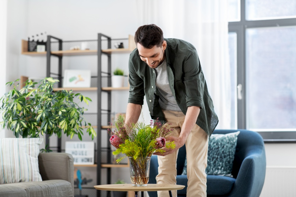 household, home improvement and interior concept - happy smiling young man placing flowers on coffee table. man placing flowers on coffee table at home
