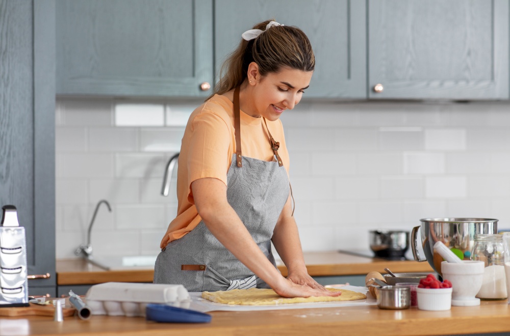 culinary, bake and people concept - happy smiling young woman cooking food on kitchen at home spreading dough on baking paper. woman cooking food and baking on kitchen at home