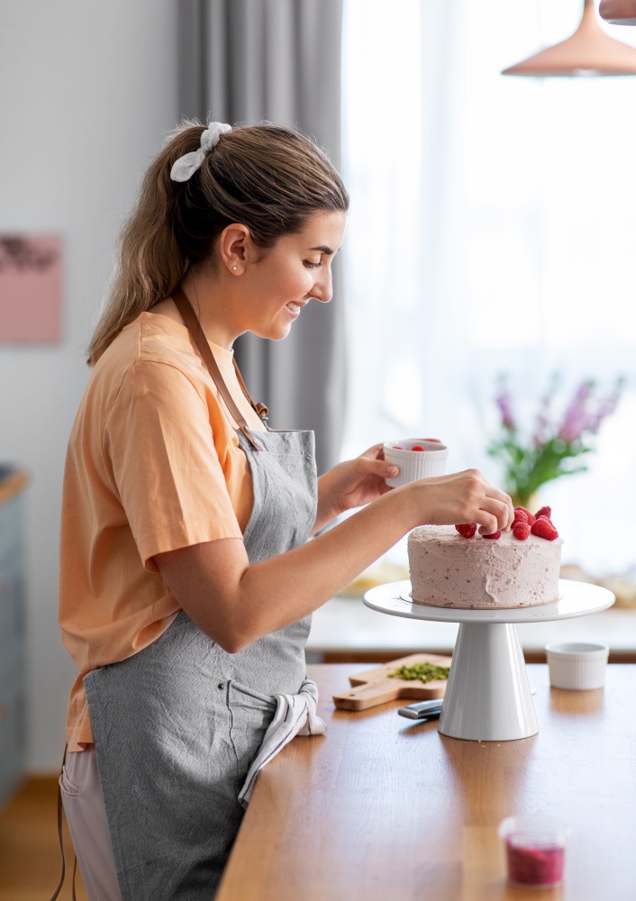 culinary, baking and cooking food concept - happy smiling young woman decorating cake with raspberries on kitchen at home. woman cooking food and baking on kitchen at home