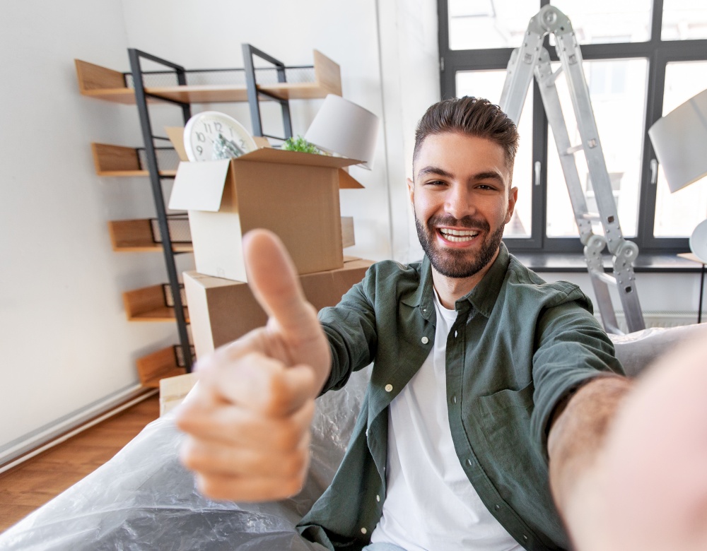 moving, people and real estate concept - happy smiling man with smartphone and boxes having video call at new home and showing thumbs up. man having video call and moving into new home