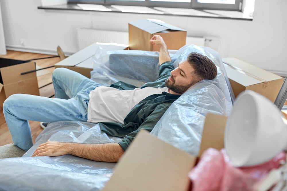 moving, people and real estate concept - tired man with boxes resting on sofa covered with plastic sheeting at new home. tired man with boxes moving to new home