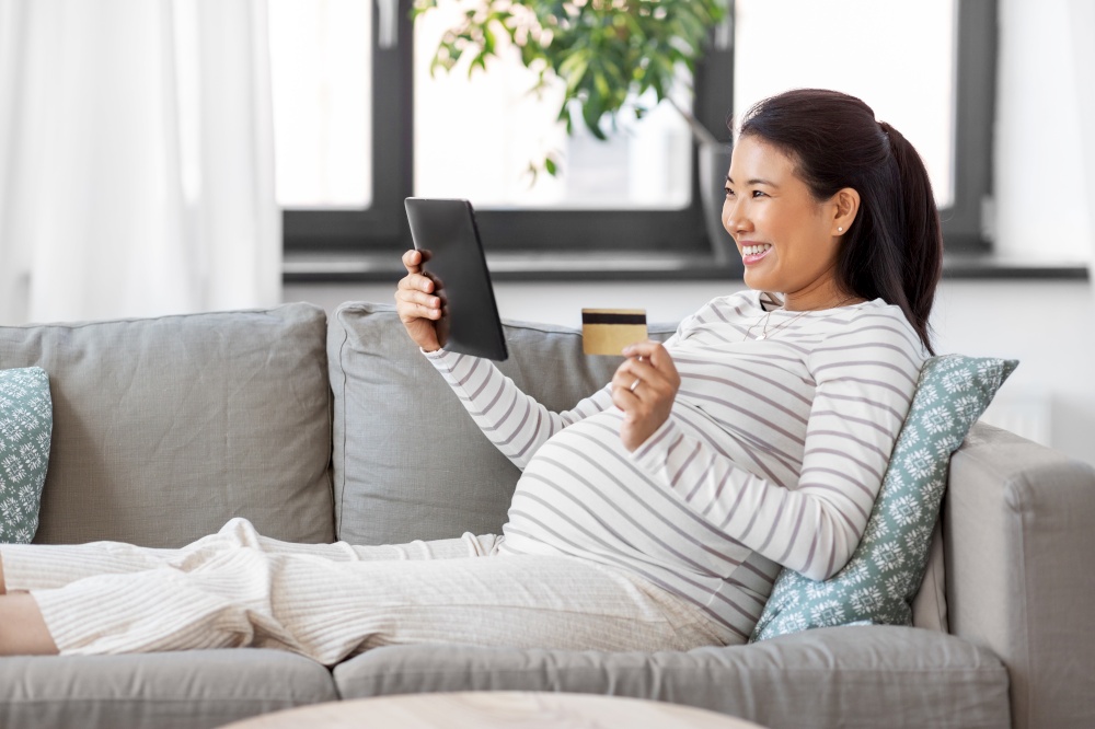 pregnancy, rest, people and expectation concept - happy smiling pregnant asian woman sitting on sofa at home. pregnant woman with tablet pc and credit card