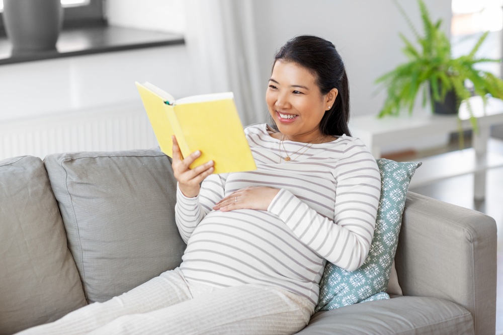 pregnancy, people and leisure concept - happy smiling pregnant asian woman sitting on sofa and reading book at home. happy pregnant woman reading book at home