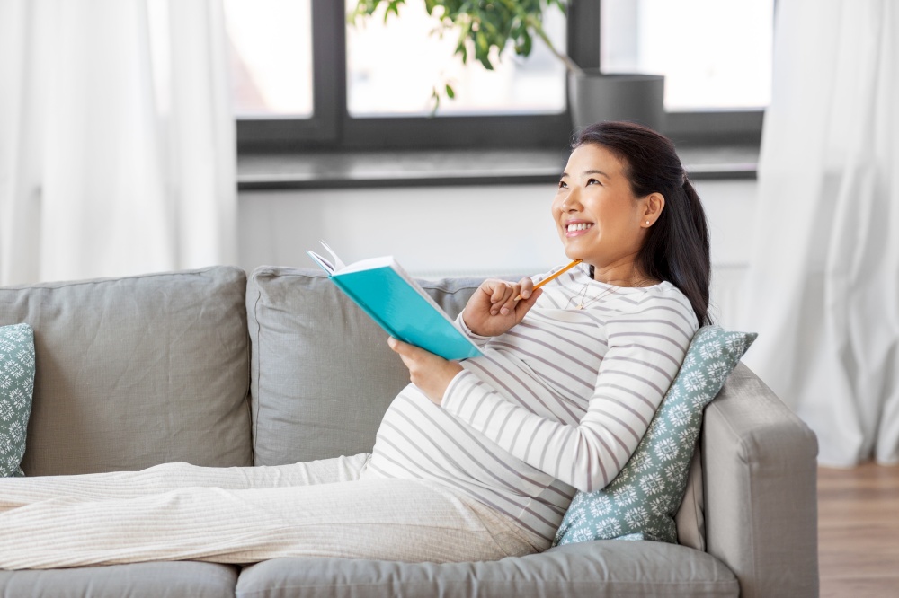pregnancy, people and leisure concept - happy smiling pregnant asian woman sitting on sofa and writing to diary or notebook at home. happy pregnant woman writing to diary at home