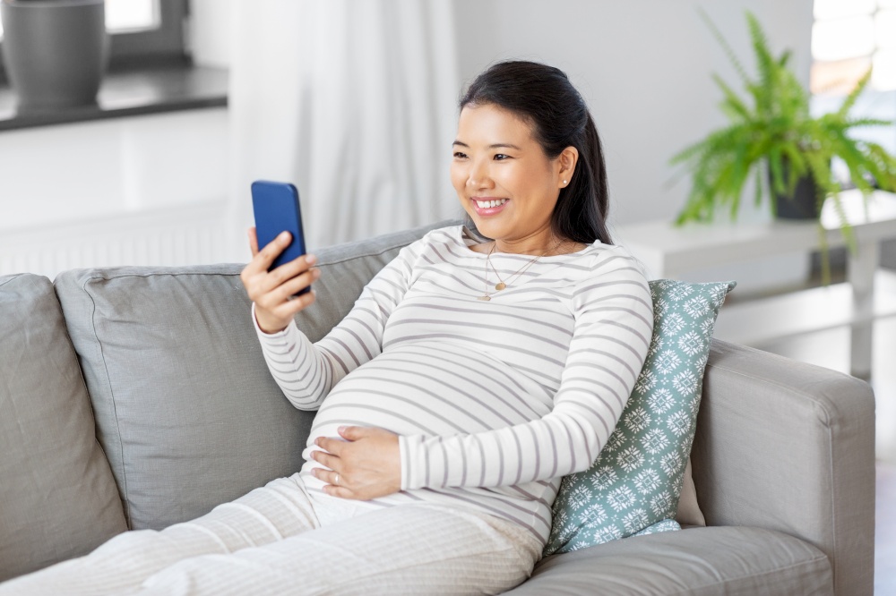 pregnancy, rest, people and expectation concept - happy smiling pregnant asian woman with smartphone sitting on sofa at home. happy pregnant woman with smartphone at home