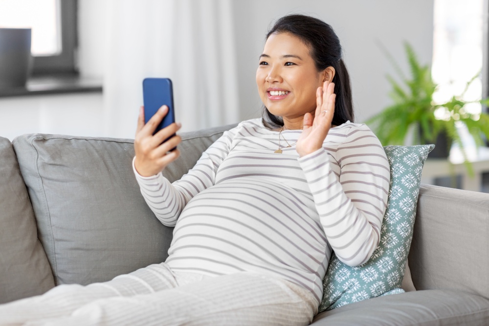 pregnancy, rest, people and expectation concept - happy smiling pregnant asian woman sitting on sofa at home having video call on smartphone. happy pregnant woman having video call on phone