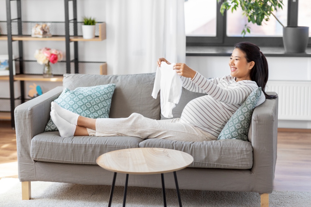 pregnancy, holidays and people concept - happy smiling pregnant asian woman with baby&rsquo;s bodysuit at home. happy pregnant woman with baby&rsquo;s bodysuit at home