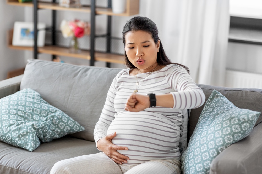 pregnancy, rest, people and expectation concept - pregnant asian woman with smart watch having labor contractions sitting on sofa at home and breathing. pregnant woman having labor contractions at home