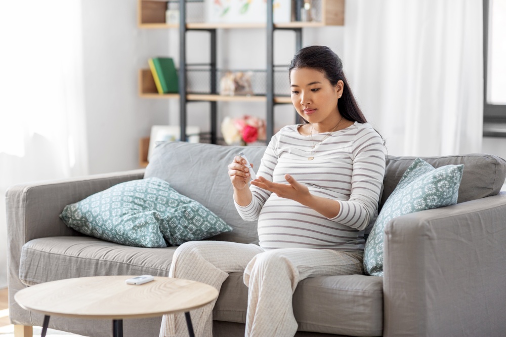 pregnancy, health and glycemia concept - pregnant asian woman checking blood sugar level with glucometer and lancing device at home. pregnant woman with glucometer making blood test