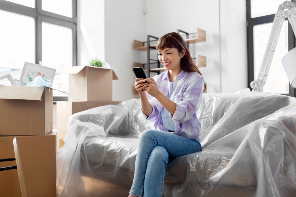 moving, people and real estate concept - happy smiling asian woman with smartphone and boxes at new home. asian woman with smartphone moving into new home