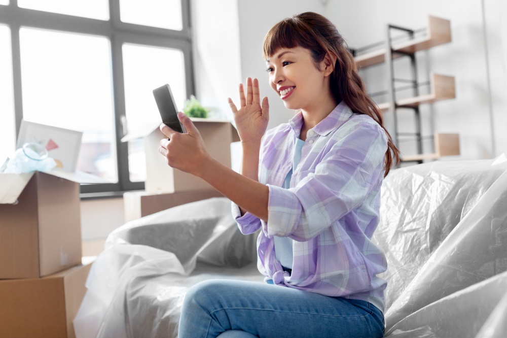 moving, people and real estate concept - happy smiling asian woman with smartphone and boxes having video call at new home. woman with phone having video call at new home