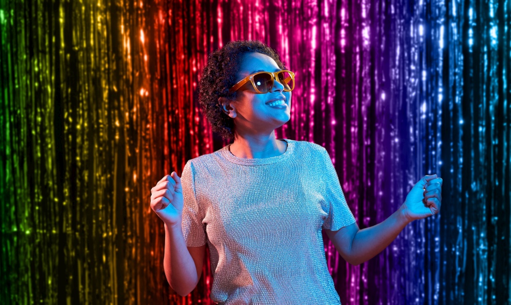 lgbt, clubbing and nightlife concept - smiling young african american woman in sunglasses dancing in ultraviolet neon lights over foil fringe curtain in rainbow colors background. african american woman dancing over neon lights