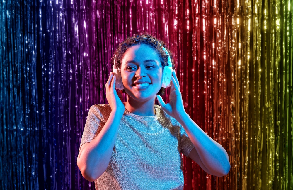 nightlife, technology and people concept - happy young african american woman in headphones listening to music at party in neon lights over rainbow foil curtain background. woman in headphones listening to music at party