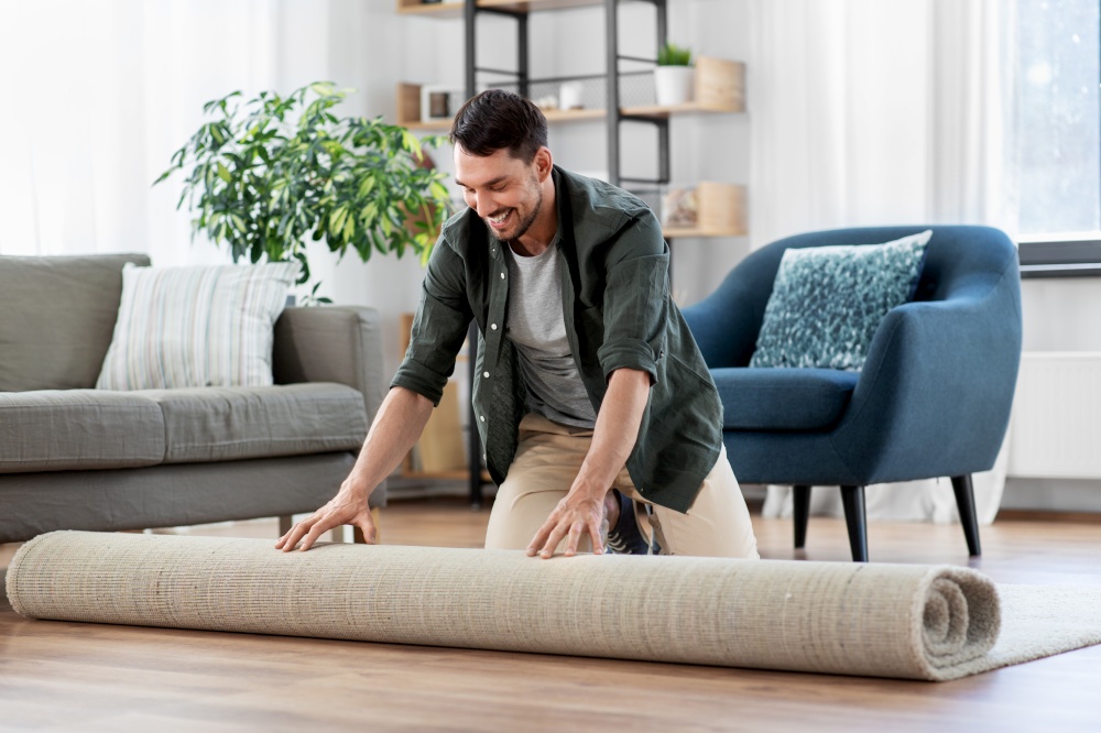 household, home improvement and interior concept - happy smiling young man unfolding carpet. happy smiling young man unfolding carpet at home