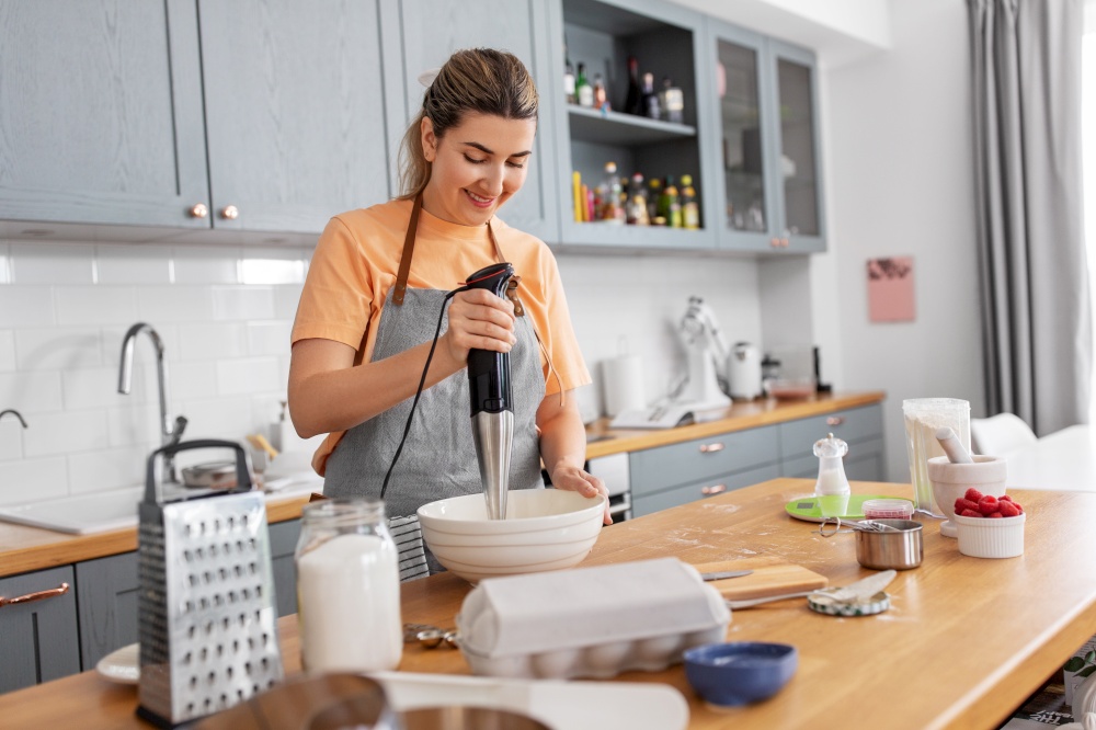 culinary, baking and people concept - happy smiling young woman cooking food on kitchen at home and using immersion blender. woman cooking food and baking on kitchen at home