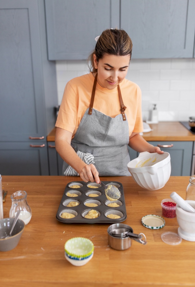 culinary, bake and people concept - happy smiling young woman cooking food on kitchen at home putting batter in baking dish for cupcakes. woman cooking food and baking on kitchen at home