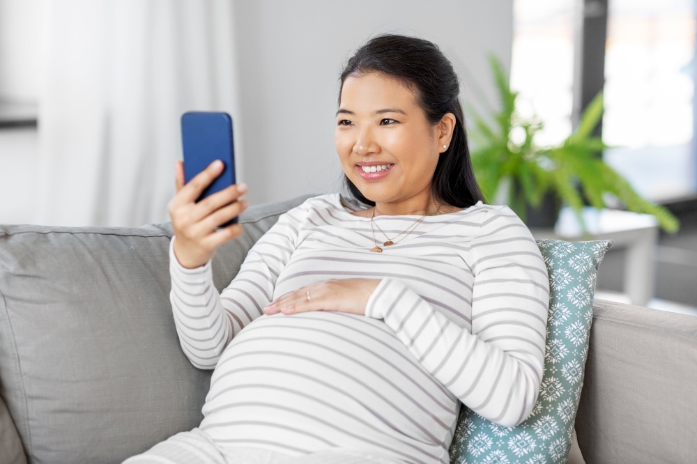 pregnancy, rest, people and expectation concept - happy smiling pregnant asian woman with smartphone sitting on sofa at home. happy pregnant woman with smartphone at home