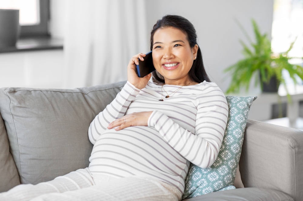 pregnancy, rest, people and expectation concept - happy smiling pregnant asian woman sitting on sofa at home and calling on smartphone. happy pregnant woman calling on smartphone at home