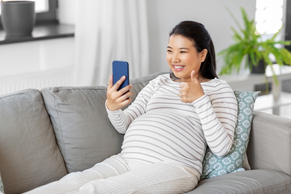 pregnancy, rest, people and expectation concept - happy smiling pregnant asian woman sitting on sofa at home having video call on smartphone and showing thumbs up. happy pregnant woman having video call on phone