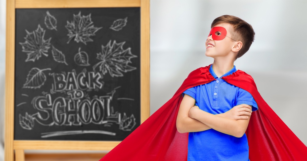 education, learning and people concept - happy boy in red super hero cape and mask over chalkboard with back to school lettering on background. boy in super hero costume over school blackboard