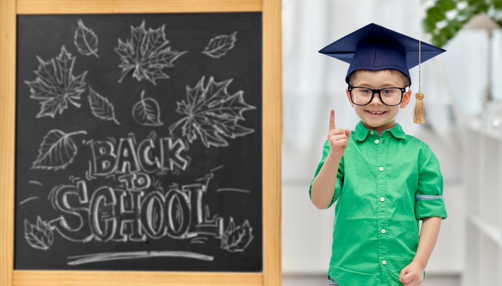 education, learning and people concept - happy little student boy in bachelor hat or mortarboard and eyeglasses pointing finger up over chalkboard with back to school lettering on background. little student boy in bachelor hat and eyeglasses