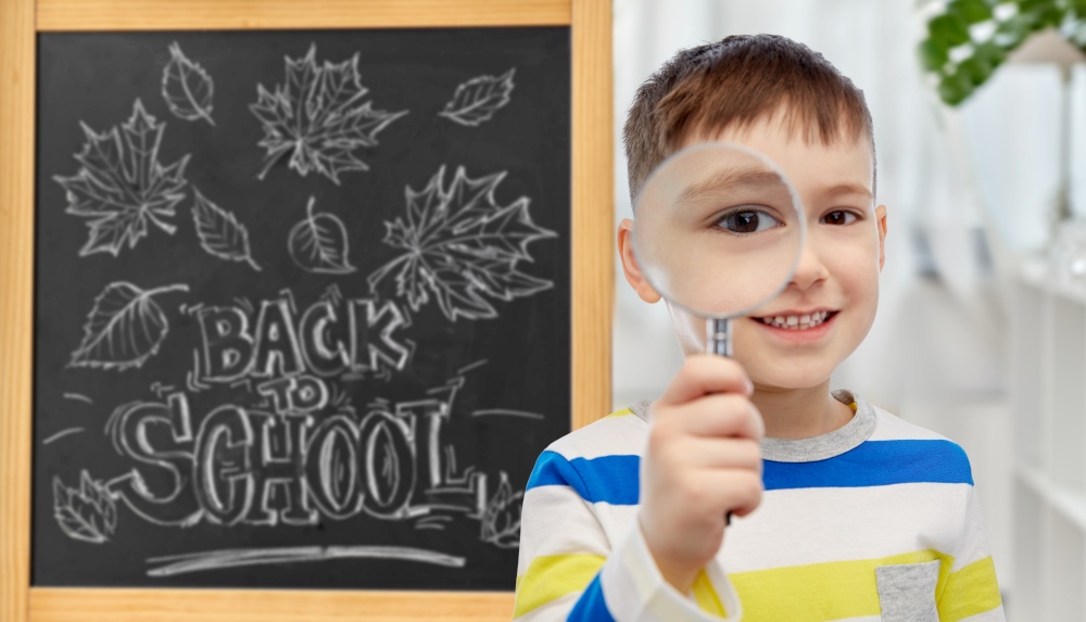 education, learning and people concept - happy little student boy looking through magnifying glass over chalkboard with back to school lettering on background. student boy looking through magnifying glass