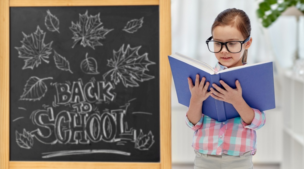 education, learning and people concept - happy little student girl in eyeglasses reading book over chalkboard with back to school lettering on background. little student girl in eyeglasses reading book