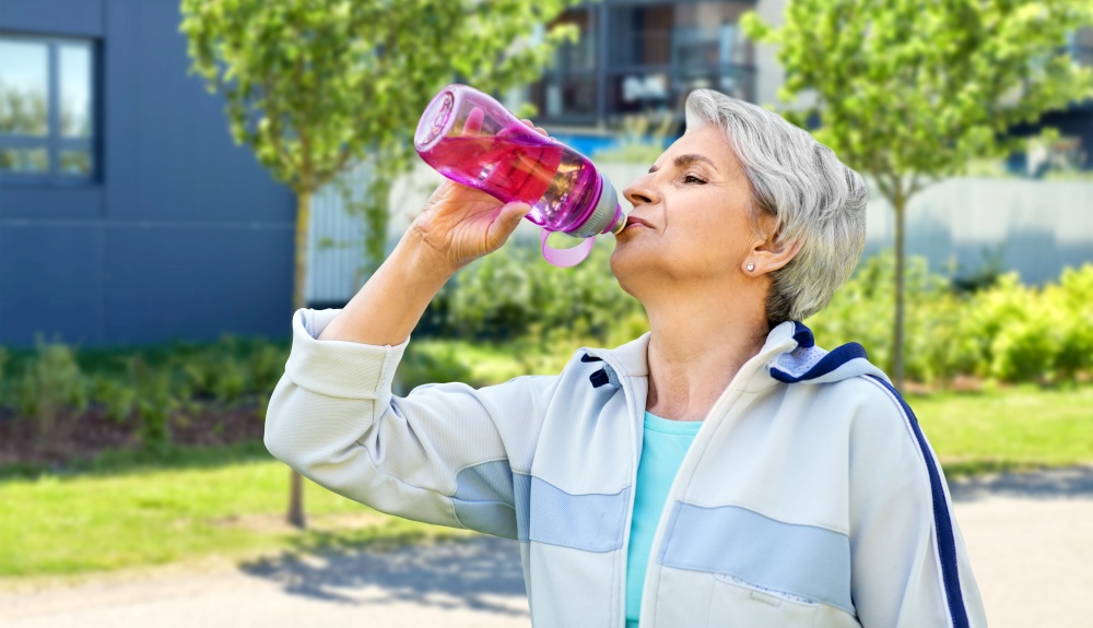 fitness, sport and healthy lifestyle concept - thirsty senior woman drinking water from bottle after exercising over city street background. old woman drinking water after exercising in city