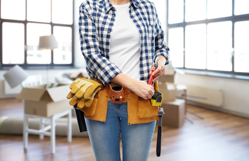 moving, repair and people concept - woman or builder with pliers and working tools on belt over new home background. woman with working tools on belt at new home