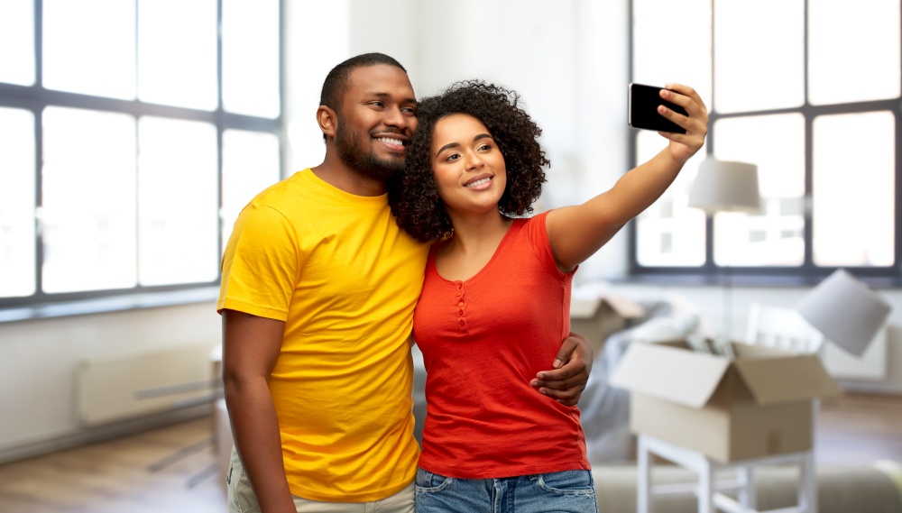 moving, real estate and people concept - happy smiling african american couple taking selfie by smartphone over new home background. african american couple taking selfie at new home