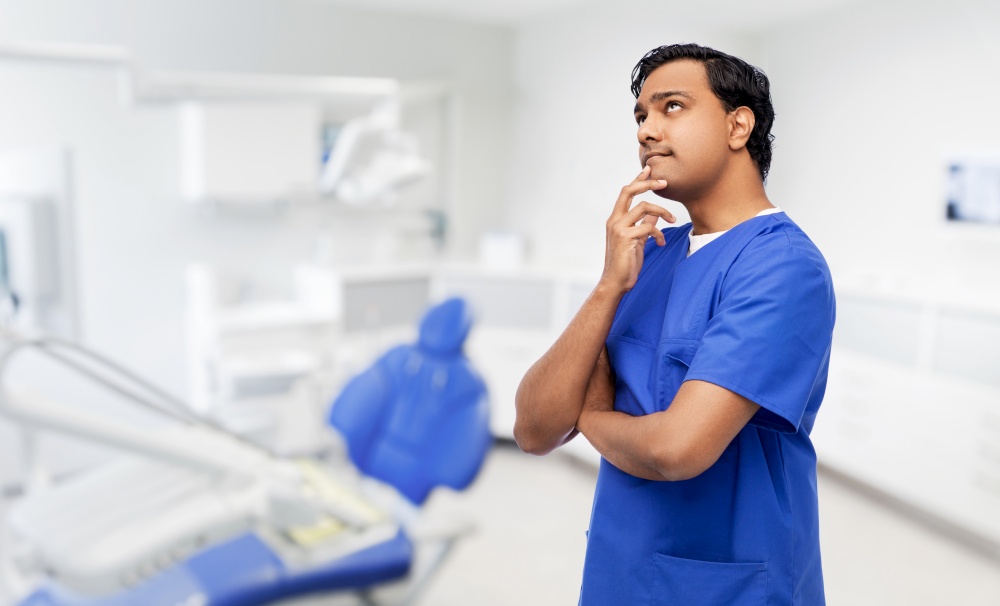 healthcare, stomatology and medicine concept - thinking indian doctor or male dentist in blue uniform over dental clinic office background. thinking indian male doctor at dental office