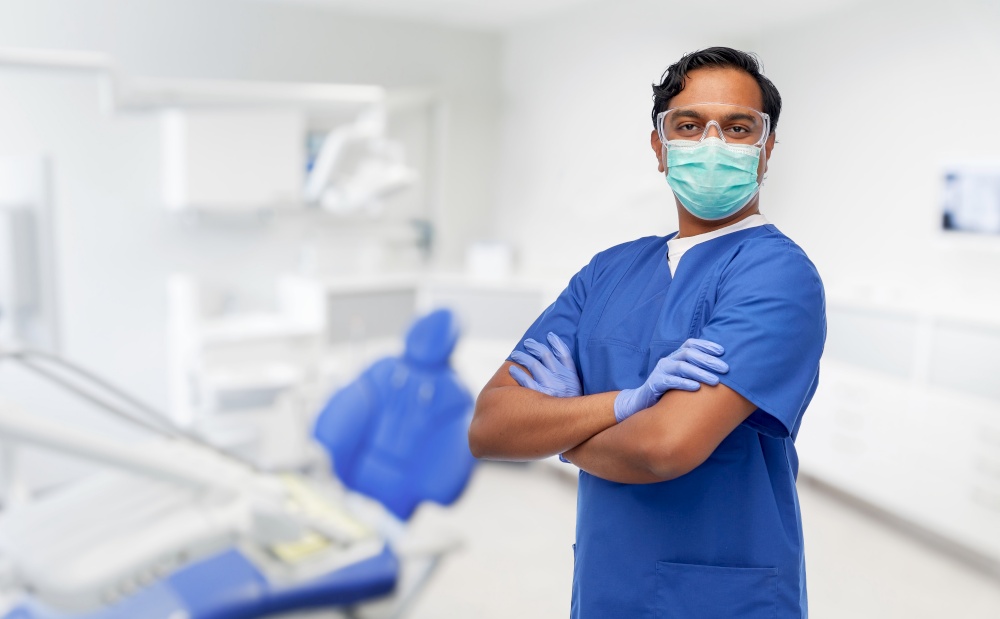 healthcare, stomatology and medicine concept - indian doctor or male dentist in blue uniform and face protective medical mask for protection from virus disease over dental clinic office background. indian male doctor in mask at dental office