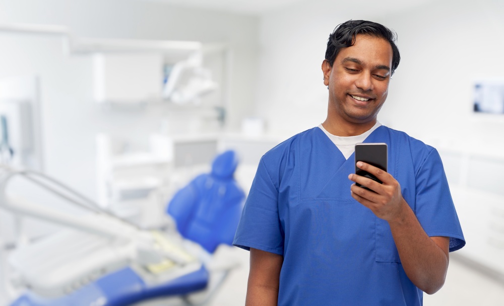 medicine, stomatology and technology concept - happy smiling indian doctor or male dentist in blue uniform using smartphone over dental clinic office background. smiling doctor using smartphone at dental office