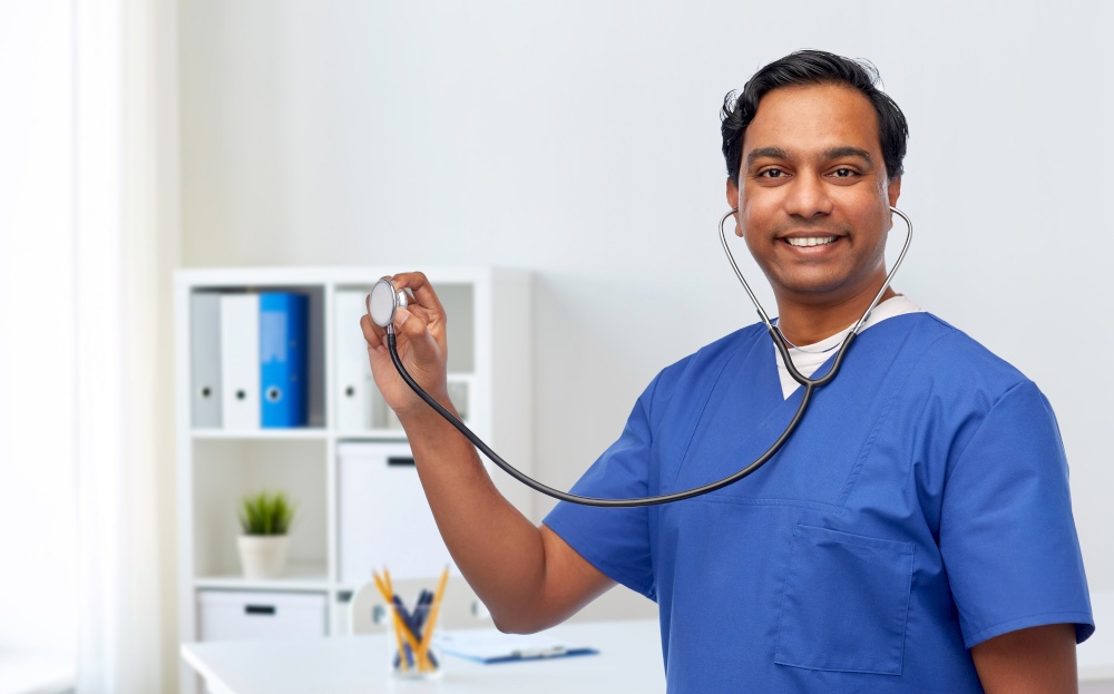 healthcare, profession and medicine concept - happy smiling indian doctor or male nurse in blue uniform with stethoscope over medical office at hospital background. happy indian male doctor or nurse with stethoscope