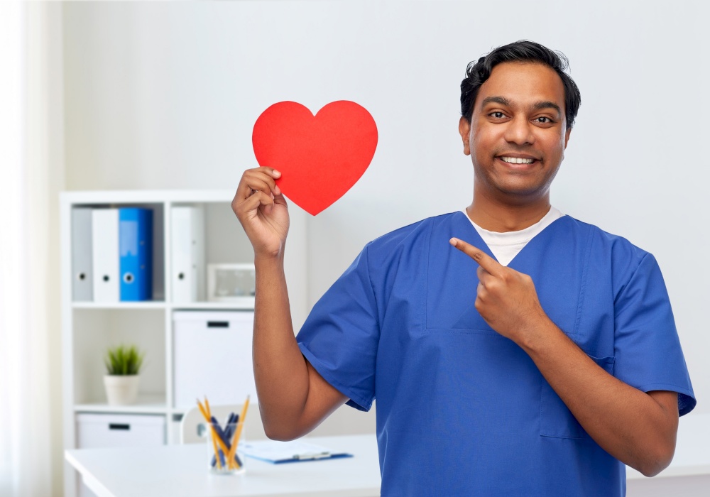 healthcare, profession and medicine concept - happy smiling indian doctor or male nurse in blue uniform with red heart over medical office at hospital background. smiling male doctor with red heart on clipboard